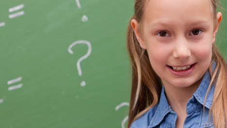 Smiling-caucasian-schoolgirl,-with-maths-questions-on-green-chalkboard