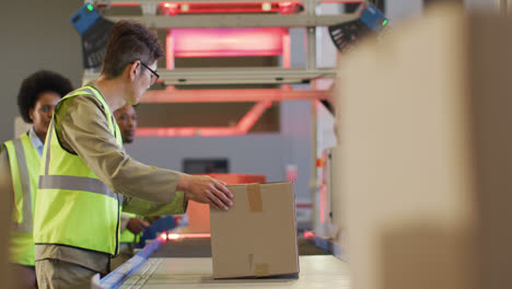 Diverse-male-and-female-workers-wearing-safety-suits-with-boxes-on-conveyor-belt-in-warehouse