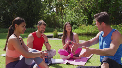 Male-yoga-instructor-talking-with-happy-diverse-group-sitting-on-mats-in-sunny-park
