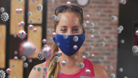 Multiple-covid-19-cells-floating-against-portrait-of-caucasian-fit-woman-wearing-a-mask-at-the-gym