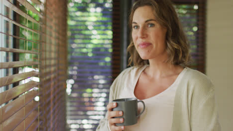 Happy-caucasian-pregnant-woman-standing-at-window-with-tea-and-touching-belly