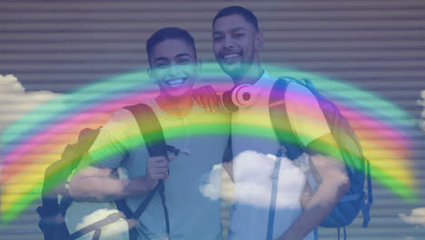 Animation-of-rainbow-and-clouds-over-smiling-gay-male-couple-with-backpacks-embracing