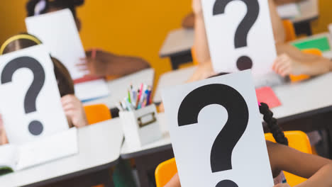 Diverse-class-of-elementary-schoolchildren-at-their-desks-holding-question-mark-signs