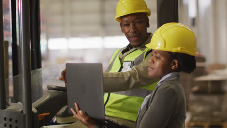 African-american-male-and-female-workers-wearing-safety-suits-and-using-laptop-in-warehouse
