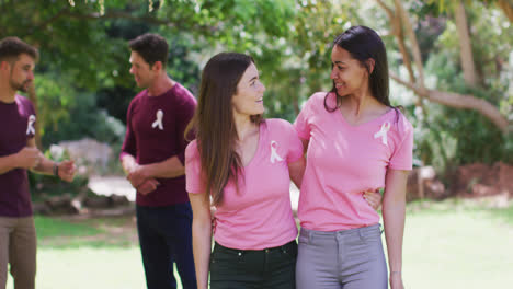 Two-happy-diverse-women-in-pink-t-shirts-and-cancer-ribbons,-embracing-and-flexing-biceps-in-park