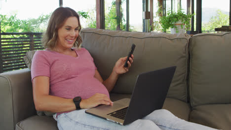 Happy-caucasian-pregnant-woman-sitting-on-sofa-and-using-smartphone-and-laptop