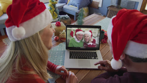 Kaukasisches-Paar-Mit-Weihnachtsmützen-Nutzt-Laptop-Für-Weihnachtsvideoanruf-Mit-Paar-Auf-Dem-Bildschirm
