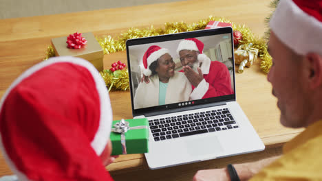 Caucasian-father-and-son-with-santa-hats-using-laptop-for-christmas-video-call-with-couple-on-screen