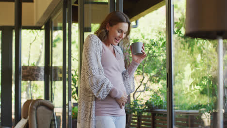 Happy-caucasian-pregnant-woman-standing-at-window-with-cup-of-tea-and-touching-belly