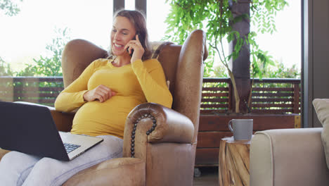 Glückliche-Kaukasische-Schwangere-Frau,-Die-Im-Sessel-Sitzt,-Telefoniert-Und-Einen-Laptop-Benutzt