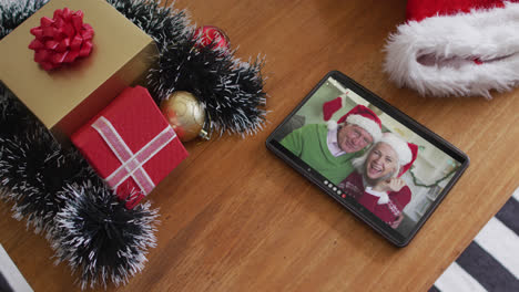 Smiling-senior-caucasian-couple-wearing-santa-hats-on-christmas-video-call-on-tablet