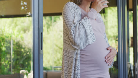 Midsection-of-caucasian-pregnant-woman-standing-at-window-with-cup-of-tea,-touching-belly