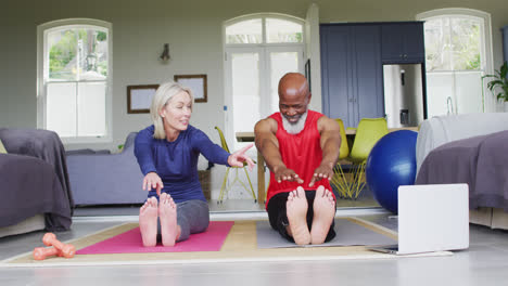 Mixed-race-senior-couple-performing-stretching-exercise-looking-at-laptop-at-home