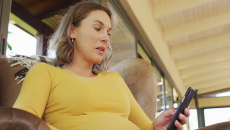 Caucasian-pregnant-woman-sitting-in-armchair-with-smartphone