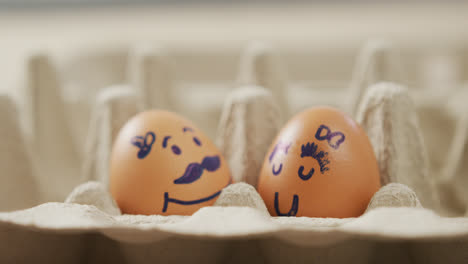 Video-Einer-Nahaufnahme-Von-Zwei-Braunen-Eiern-Mit-Gezeichneten-Gesichtern-Im-Eierkarton-Hintergrund