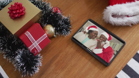 Smiling-senior-african-american-couple-wearing-santa-hats-on-christmas-video-call-on-tablet