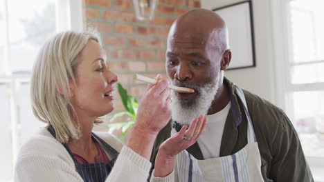 Mixed-race-senior-couple-wearing-aprons-tasting-food-while-cooking-in-the-kitchen-at-home