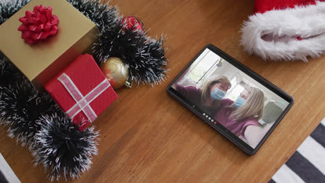 Smiling-caucasian-mother-and-daughter-with-face-masks-on-christmas-video-call-on-tablet