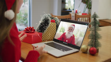 Caucasian-woman-on-laptop-video-call-with-santa-claus-at-christmas-time
