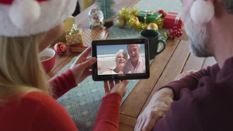 Kaukasisches-Paar-Mit-Weihnachtsmützen-Nutzt-Tablet-Für-Weihnachtsvideoanruf-Mit-Paar-Auf-Dem-Bildschirm