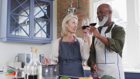 Älteres-Paar-Mit-Gemischter-Abstammung-Trägt-Schürzen-Und-Trinkt-Wein-Beim-Kochen-In-Der-Küche-Zu-Hause