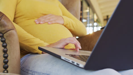 Midsection-of-caucasian-pregnant-woman-sitting-in-armchair-with-laptop