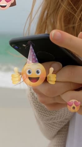 Animation-of-emoticons-over-hands-of-caucasian-woman-using-smartphone-at-beach