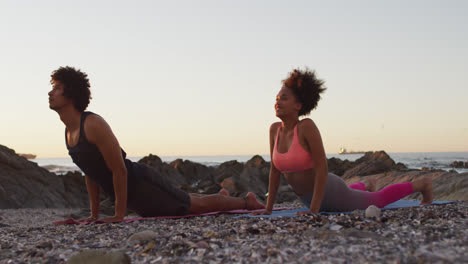 African-american-couple-practicing-yoga-together-on-rocks-near-the-sea-during-sunset