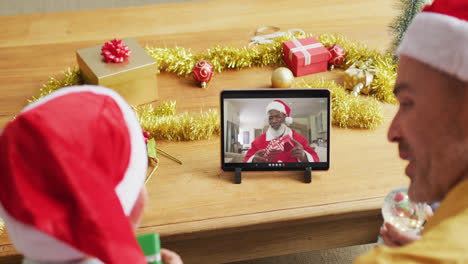 Kaukasischer-Vater-Und-Sohn-Mit-Weihnachtsmützen,-Die-Ein-Tablet-Für-Einen-Weihnachtsvideoanruf-Mit-Dem-Weihnachtsmann-Auf-Dem-Bildschirm-Nutzen