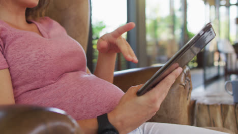 Midsection-of-caucasian-pregnant-woman-sitting-in-armchair-using-tablet