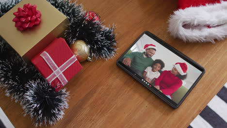 Smiling-african-american-family-wearing-santa-hats-on-christmas-video-call-on-tablet