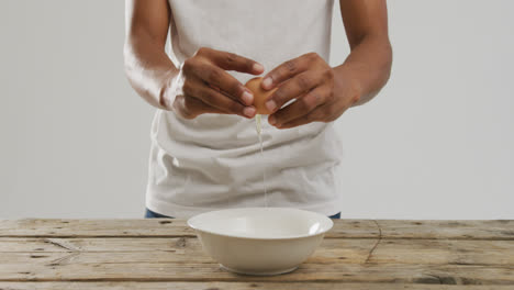 Video-of-biracial-man-cracking-egg-into-white-bowl-on-wooden-surface-and-white-background