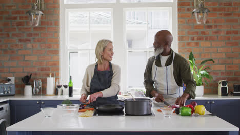 Mixed-race-senior-couple-wearing-aprons-cooking-food-together-in-the-kitchen-at-home