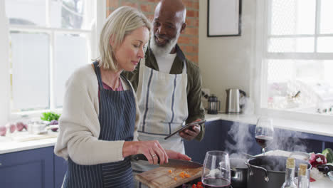 Älteres-Paar-Mit-Gemischter-Abstammung-Trägt-Schürzen-Und-Benutzt-Ein-Digitales-Tablet-Beim-Kochen-In-Der-Küche-Zu-Hause