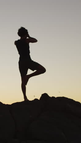 Silhouetted-young-biracial-man-practices-yoga-outdoors-at-sunset