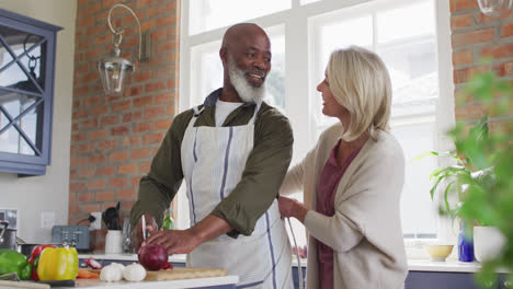 Caucasian-senior-woman-tying-apron-to-her-husband-in-the-kitchen-at-home