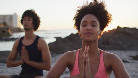 African-american-couple-practicing-yoga-and-meditating-together-on-rocks-near-the-sea-during-sunset