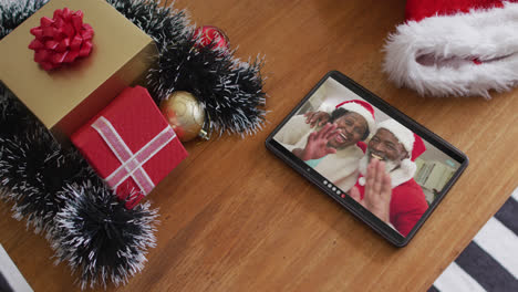 Smiling-senior-african-american-couple-wearing-santa-hats-on-christmas-video-call-on-tablet