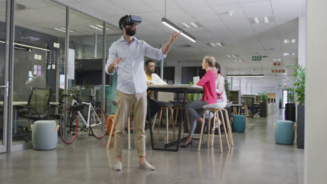 Happy-biracial-businessman-using-vr-headset-over-colleagues-in-office