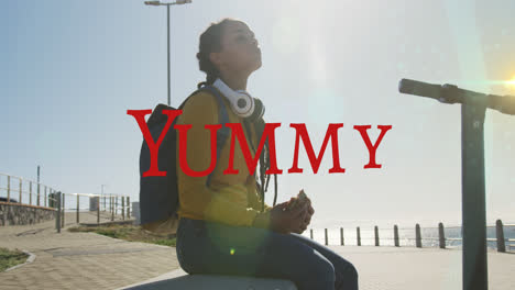 Animation-of-yummy-text-over-biracial-woman-with-scooter-eating-on-promenade