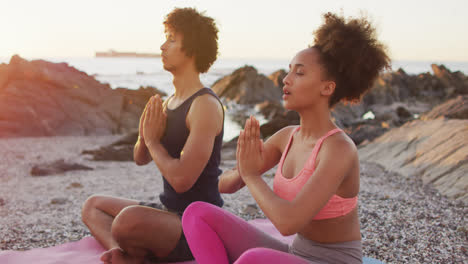 African-american-couple-practicing-yoga-and-meditating-together-on-rocks-near-the-sea-during-sunset