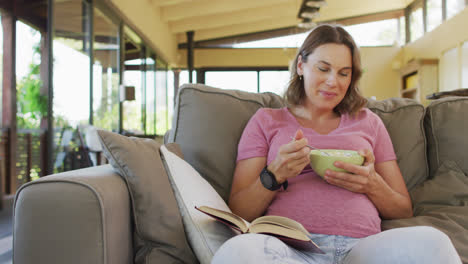 Relaxed-caucasian-pregnant-woman-lying-on-sofa,-eating-and-reading-book