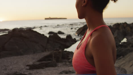 Porträt-Einer-Afroamerikanischen-Frau-Mit-Yogamatte,-Die-Bei-Sonnenuntergang-Auf-Den-Felsen-In-Der-Nähe-Des-Meeres-Steht