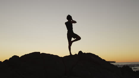 Silhouette-Eines-Afroamerikanischen-Mannes,-Der-Bei-Sonnenuntergang-Yoga-Praktiziert-Und-Auf-Felsen-In-Der-Nähe-Des-Meeres-Meditiert