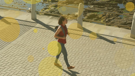 Animation-of-yellow-spots-over-biracial-woman-walking-on-promenade