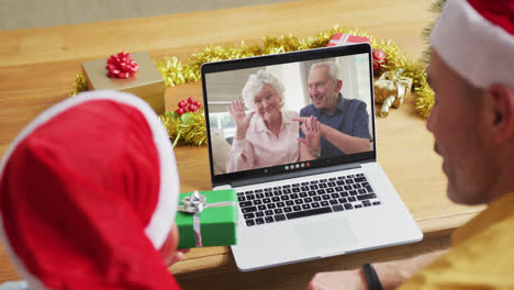 Kaukasischer-Vater-Und-Sohn-Mit-Weihnachtsmützen-Nutzen-Laptop-Für-Weihnachtsvideoanruf-Mit-Paar-Auf-Dem-Bildschirm