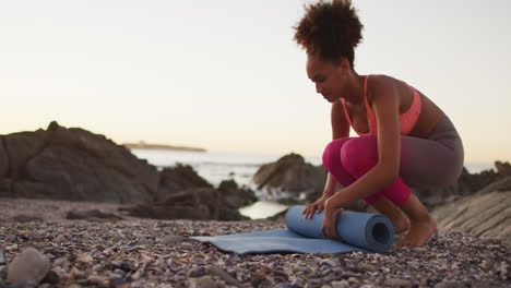 Afroamerikanische-Frau-Rollt-Ihre-Yogamatte-Bei-Sonnenuntergang-Auf-Den-Felsen-In-Der-Nähe-Des-Meeres