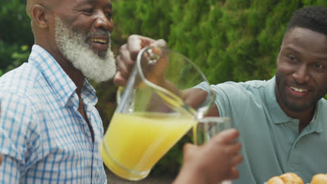 Happy-african-american-family-pouring-juice-and-having-breakfast-in-garden