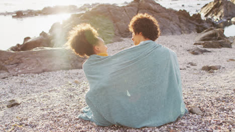 Rear-view-of-african-american-couple-wrapped-in-blanket-sitting-on-the-rocks-near-the-sea