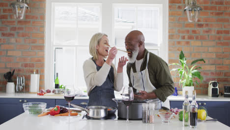 Älteres-Paar-Mit-Gemischter-Abstammung-Trägt-Schürzen-Und-Probiert-Essen-Beim-Kochen-In-Der-Küche-Zu-Hause
